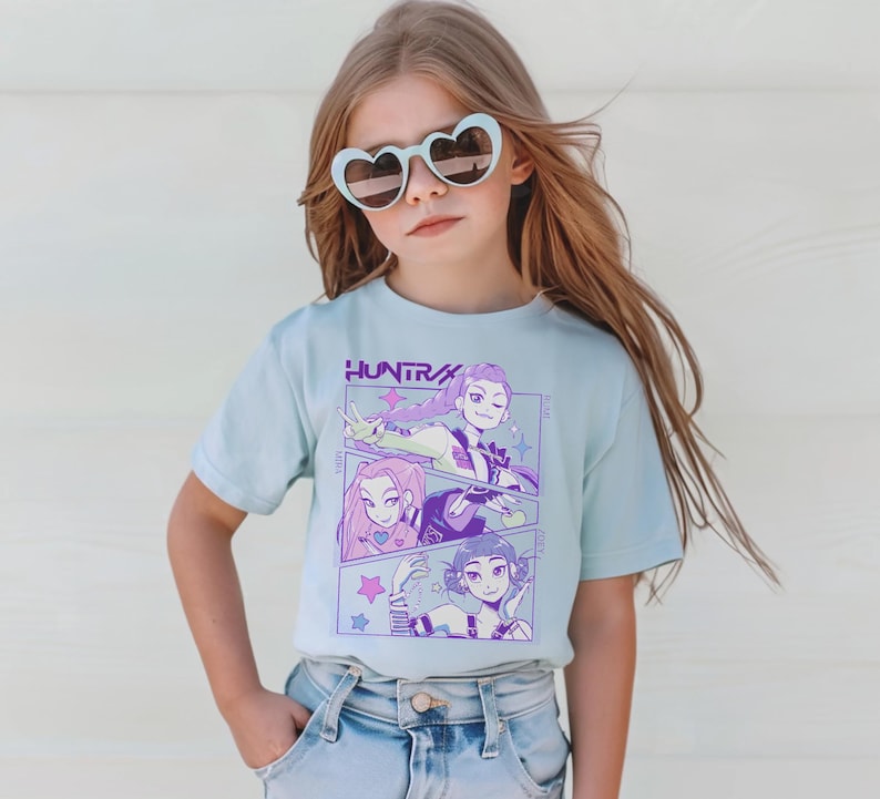Peut inclure: Un t-shirt bleu clair avec un graphisme de trois personnages de style anime aux accents violets et roses. Le mot "HUNTRA" est imprim&eacute; au-dessus du graphisme en violet. Le t-shirt est port&eacute; par une jeune personne portant des lunettes de soleil en forme de c&oelig;ur.