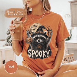 Pu&ograve; includere: Una t-shirt arancione con la grafica di un procione che indossa un cappello da strega e un mantello, mentre tiene in mano una zucca e un bicchiere. Il procione &egrave; davanti a una luna piena e alberi spogli, con la scritta SPOOKY in bianco sotto.