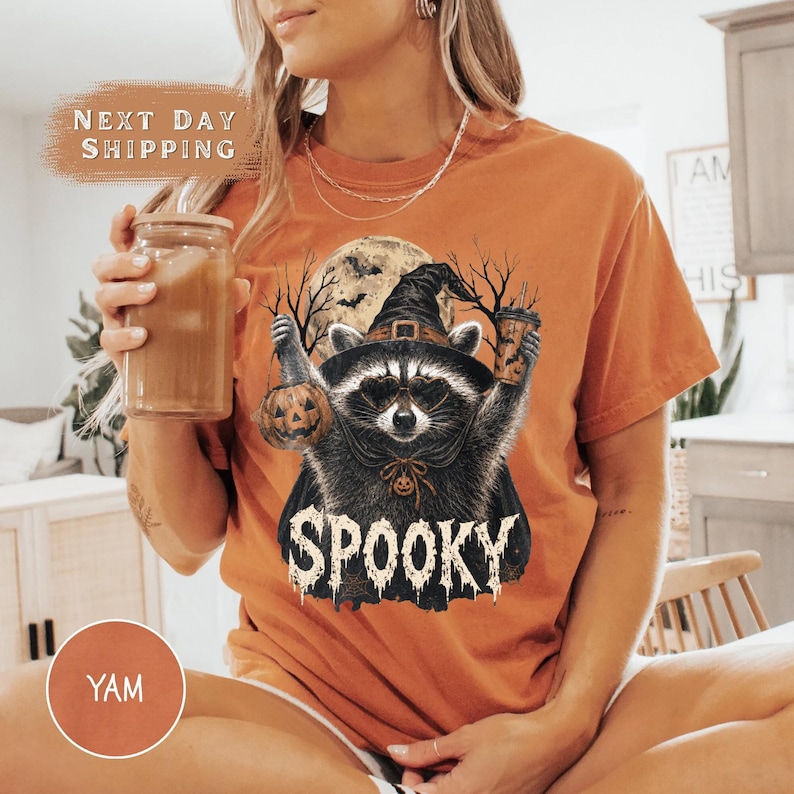 Pu&ograve; includere: Una t-shirt arancione con la grafica di un procione che indossa un cappello da strega e un mantello, mentre tiene in mano una zucca e un bicchiere. Il procione &egrave; davanti a una luna piena e alberi spogli, con la scritta SPOOKY in bianco sotto.