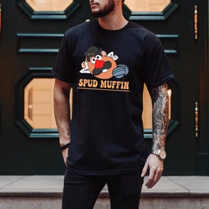 May include: A black t-shirt with a cartoon image of Mr. Potato Head wearing a baseball cap and holding a baseball bat. The text "SPUD MUFFIN" is printed in orange below the image.