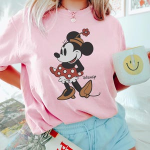 May include: Pink t-shirt with a vintage cartoon image of Minnie Mouse wearing a red polka dot dress and the word "DISNEY" printed on the bottom right corner.