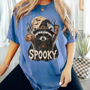 Pu&ograve; includere: Una t-shirt blu a maniche corte con la grafica di un procione che indossa un cappello da strega e occhiali, che tiene in mano un secchiello a forma di zucca e una bevanda. Il procione &egrave; davanti a una luna piena e alberi, con la scritta SPOOKY.