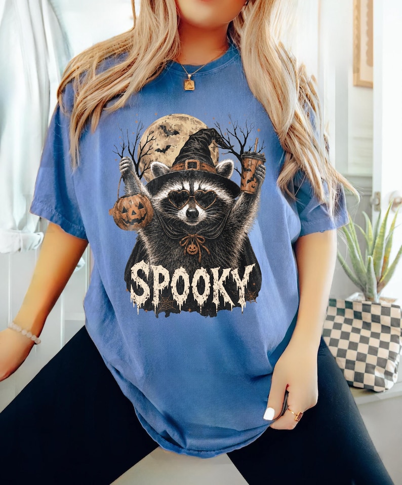 Pu&ograve; includere: Una t-shirt blu a maniche corte con la grafica di un procione che indossa un cappello da strega e occhiali, che tiene in mano un secchiello a forma di zucca e una bevanda. Il procione &egrave; davanti a una luna piena e alberi, con la scritta SPOOKY.