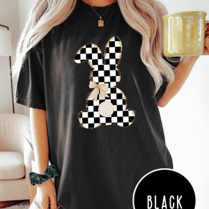 Op de afbeelding: Zwart T-shirt met een geruite konijnenprint. Het konijn heeft een gouden strik en is omrand met goud. De tekst "BLACK" is zichtbaar in de rechterbenedenhoek van de afbeelding.
