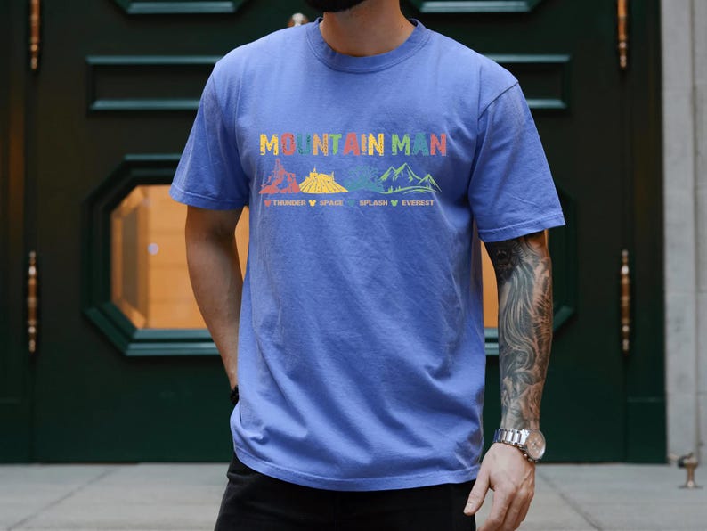 May include: A blue t-shirt with the text "Mountain Man" printed on the front. The text is surrounded by colorful illustrations of mountains with the names "Thunder", "Space", "Splash", and "Everest" printed below.