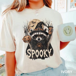 Pu&ograve; includere: Una t-shirt color avorio con la grafica di un procione che indossa un cappello da strega e occhiali da sole a forma di cuore. Il procione tiene un secchiello a forma di zucca e un bicchiere. Sullo sfondo una luna piena e alberi spogli.