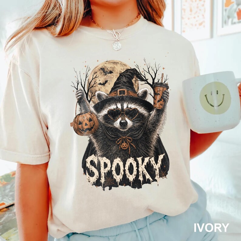 Pu&ograve; includere: Una t-shirt color avorio con la grafica di un procione che indossa un cappello da strega e occhiali da sole a forma di cuore. Il procione tiene un secchiello a forma di zucca e un bicchiere. Sullo sfondo una luna piena e alberi spogli.
