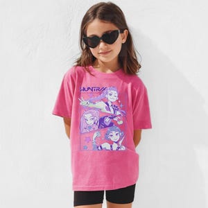 Peut inclure: Un t-shirt rose avec le mot "HUNTR" imprim&eacute; dessus. Le t-shirt pr&eacute;sente un graphisme de trois personnages de style anime dans un design de bande dessin&eacute;e. La personne sur l'image porte des lunettes de soleil en forme de c&oelig;ur et un short noir.