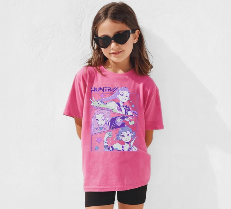 Peut inclure: Un t-shirt rose avec le mot "HUNTR" imprim&eacute; dessus. Le t-shirt pr&eacute;sente un graphisme de trois personnages de style anime dans un design de bande dessin&eacute;e. La personne sur l'image porte des lunettes de soleil en forme de c&oelig;ur et un short noir.