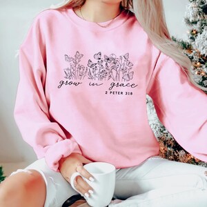 May include: A light pink sweatshirt with a floral and butterfly design, and the words "grow in grace" and "2 Peter 3:18" in black script. The sweatshirt is a crew neck style.
