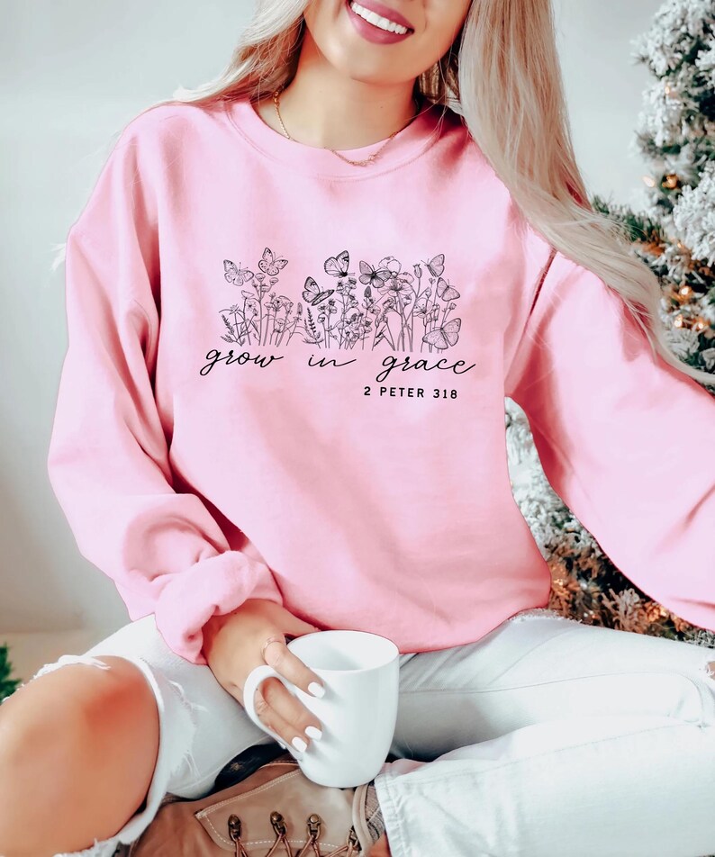 May include: A light pink sweatshirt with a floral and butterfly design, and the words "grow in grace" and "2 Peter 3:18" in black script. The sweatshirt is a crew neck style.