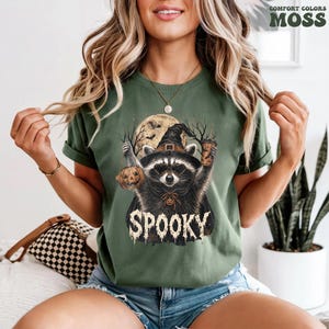 Pu&ograve; includere: Una t-shirt verde muschio con la grafica di un procione che indossa un cappello da strega, tiene un secchiello a forma di zucca e una bevanda. Il procione &egrave; davanti a una luna piena con rami spogli. La parola SPOOKY &egrave; stampata in bianco sotto.