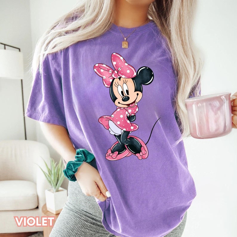 May include: Purple t-shirt featuring a graphic of Minnie Mouse wearing a pink polka dot dress and a red bow.