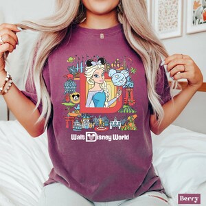 Może przedstawiać: Fioletowy T-shirt z grafiką przedstawiającą animowaną Elsę z Krainy Lodu. Grafika zawiera czerwoną literę D z r&oacute;żnymi atrakcjami i postaciami z Disney World. Pod grafiką znajduje się napis "Walt Disney World".