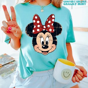 May include: A chalky mint short-sleeve t-shirt featuring a distressed graphic of Minnie Mouse with a red and white polka dot bow. The shirt includes the text COMFORT COLORS CHALKY MINT in the upper right corner. The design is printed on a crew neck shirt.