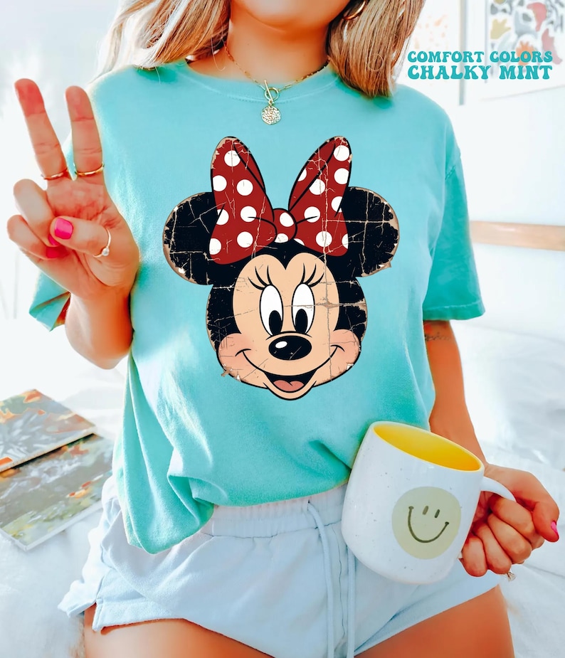 May include: A chalky mint short-sleeve t-shirt featuring a distressed graphic of Minnie Mouse with a red and white polka dot bow. The shirt includes the text COMFORT COLORS CHALKY MINT in the upper right corner. The design is printed on a crew neck shirt.