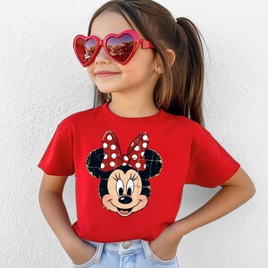 May include: A bright red short-sleeved t-shirt featuring a distressed graphic print of Minnie Mouse with her signature polka dot bow. The shirt is paired with light blue denim jeans and red heart-shaped sunglasses with dark tinted lenses.