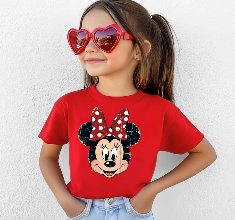 May include: A bright red short-sleeved t-shirt featuring a distressed graphic print of Minnie Mouse with her signature polka dot bow. The shirt is paired with light blue denim jeans and red heart-shaped sunglasses with dark tinted lenses.