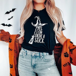 Pu&ograve; includere: T-shirt nero con cappello da strega bianco e grafica scopa. Il testo recita "YES I CAN DRIVE A STICK." Questo design a tema Halloween &egrave; perfetto per la stagione spettrale. Ritagli decorativi di pipistrelli neri sono sullo sfondo.