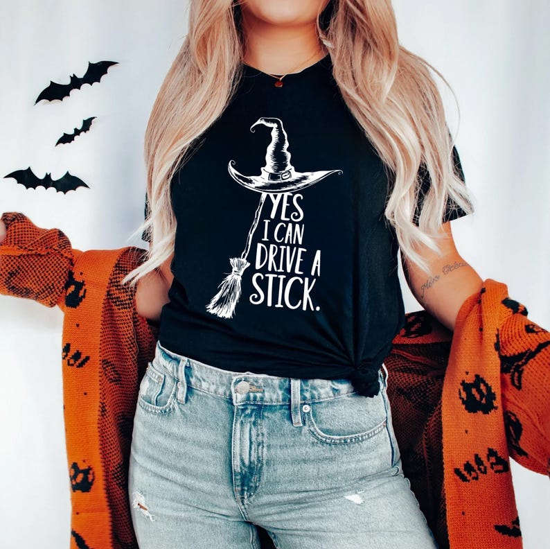 Pu&ograve; includere: T-shirt nero con cappello da strega bianco e grafica scopa. Il testo recita "YES I CAN DRIVE A STICK." Questo design a tema Halloween &egrave; perfetto per la stagione spettrale. Ritagli decorativi di pipistrelli neri sono sullo sfondo.