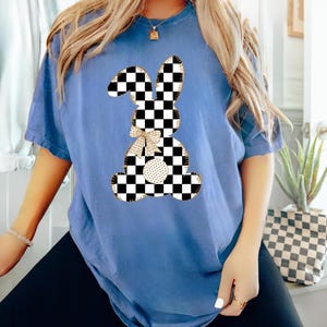 Op de afbeelding: Een blauw T-shirt met een geruite konijnenprint. Het konijn is omrand met goud en heeft een gouden strik.