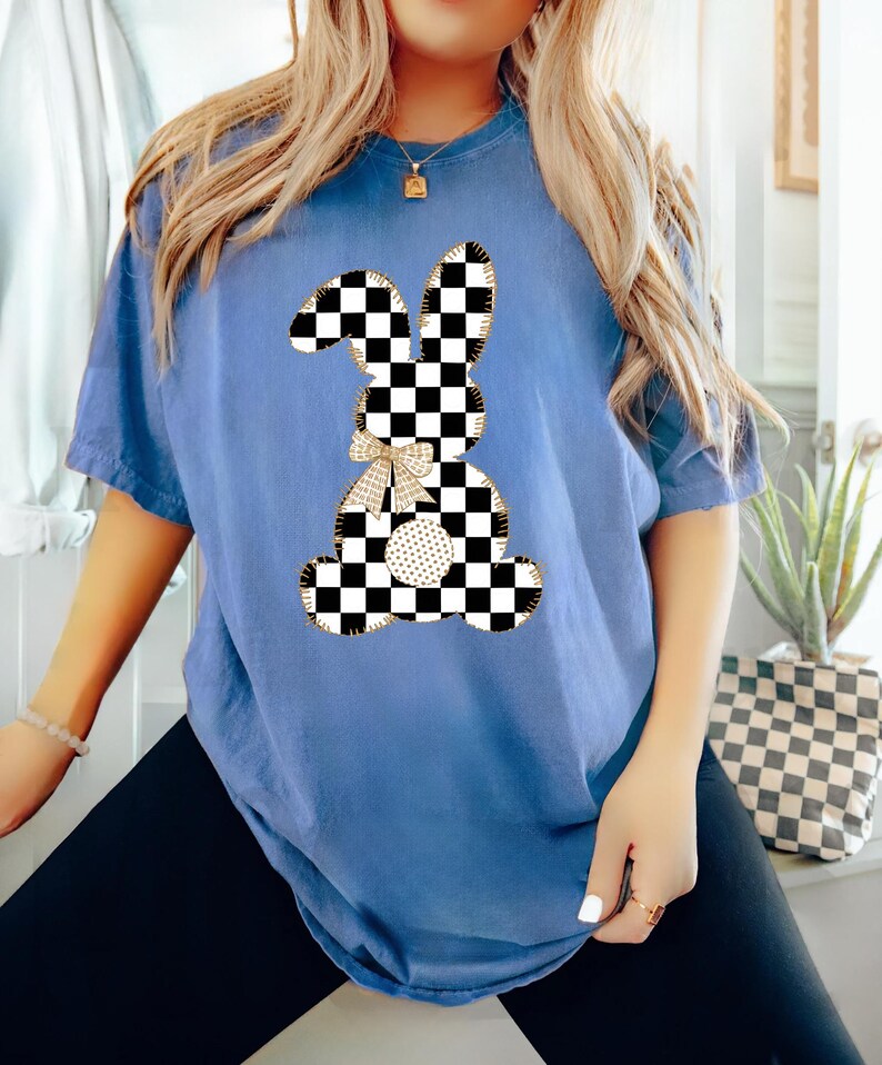 Op de afbeelding: Een blauw T-shirt met een geruite konijnenprint. Het konijn is omrand met goud en heeft een gouden strik.
