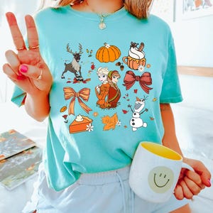 May include: A light blue t-shirt featuring a variety of fall-themed graphics. The design includes a reindeer, pumpkin, cupcake, bows, pie, and characters from a popular animated movie. A white mug with a yellow interior and a smiley face is also visible.