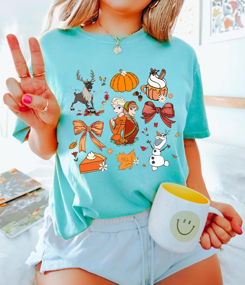 May include: A light blue t-shirt featuring a variety of fall-themed graphics. The design includes a reindeer, pumpkin, cupcake, bows, pie, and characters from a popular animated movie. A white mug with a yellow interior and a smiley face is also visible.
