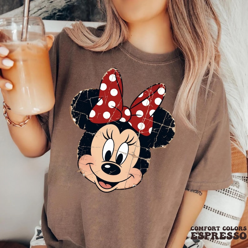 May include: A brown short-sleeve t-shirt featuring a distressed graphic of a cartoon mouse head with a red and white polka dot bow. The shirt has a crew neckline and a relaxed fit, showcasing a vintage-style print with visible cracks.