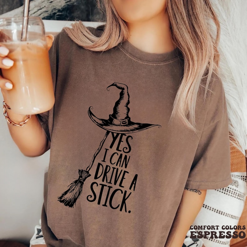Pu&ograve; includere: T-shirt color espresso con cappello da strega nero, scopa e la scritta "YES I CAN DRIVE A STICK." La maglietta &egrave; realizzata in materiale Comfort Colors.
