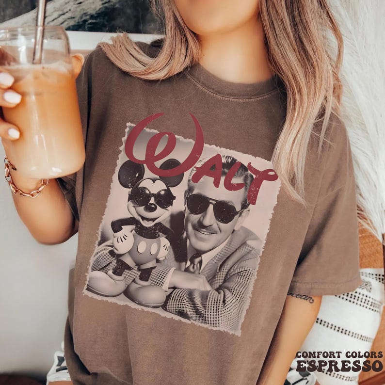 Op de afbeelding: Een bruin t-shirt met korte mouwen en een grafische print van een zwart-witfoto van Mickey Mouse en Walt Disney, beiden met een zonnebril op. Het woord Walt staat in grote rode letters in schrijfschrift boven de afbeelding gedrukt.