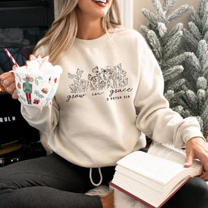 May include: Cream-colored sweatshirt with a floral and butterfly design, and the words "grow in grace" and "2 Peter 3:18" in black script. The sweatshirt has a crew neck and long sleeves. A mug with a festive design is held in the left hand.