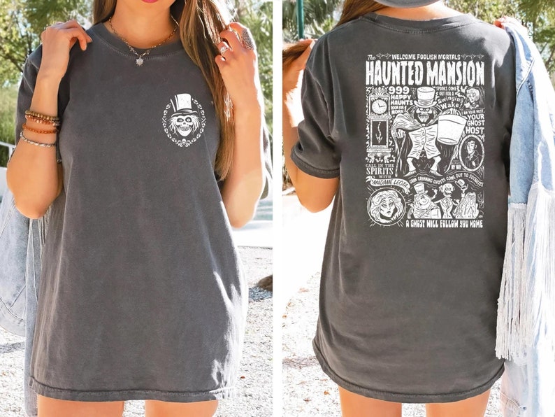 May include: A dark gray t-shirt with a white graphic print of the Haunted Mansion attraction at Disney World. The front of the shirt features a ghost with a top hat. The back of the shirt features a detailed illustration of the Haunted Mansion with text that reads "Welcome Foolish Mortals Haunted Mansion 999 Haunts Your Ghost Host Call on the Spirits in the Mansion A Ghost Will Follow You Home."
