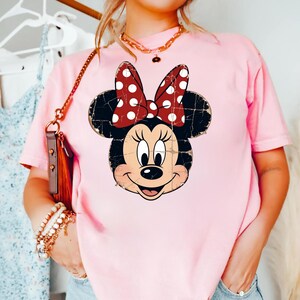 May include: A light pink short-sleeved t-shirt featuring a distressed graphic print of a cartoon mouse face with large round ears and a red bow with white polka dots centred on the chest. The graphic has a weathered, vintage-style texture.
