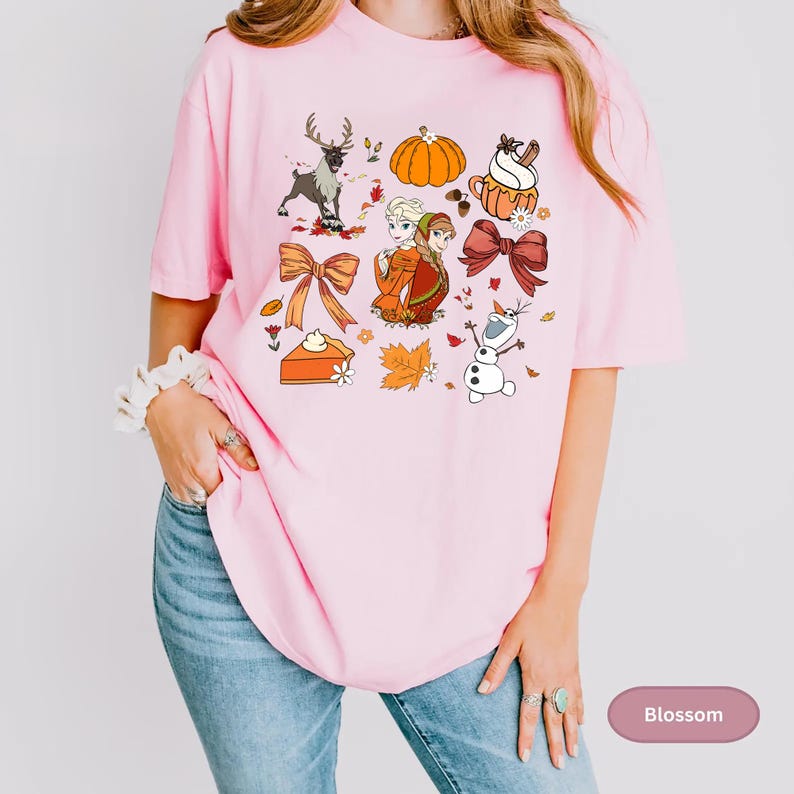 May include: A light pink t-shirt featuring a collection of fall-themed illustrations. The design includes a pumpkin, reindeer, pie, and characters from a popular animated film. The shirt is a relaxed fit.