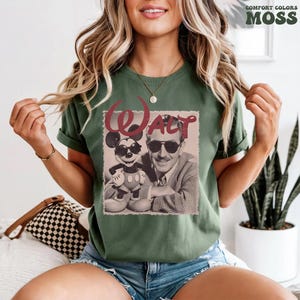 Op de afbeelding: Een mosgroen t-shirt met een grafische print van Walt Disney en Mickey Mouse die allebei een zonnebril dragen. De tekst "Walt" is in een groot, gestileerd kastanjebruin lettertype boven de figuren gedrukt.