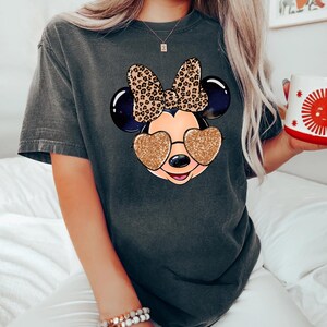 以下が含まれることがあります： レオパード柄のリボンとキラキラのハート型のサングラスをかけたマウスの漫画の画像がプリントされたダークグレーのTシャツ。