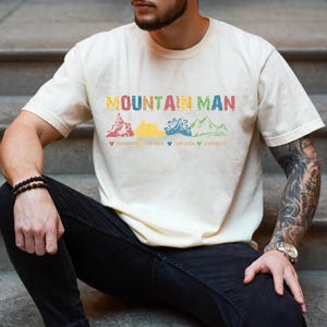 May include: A person wearing a light beige t-shirt with the text "Mountain Man" and a graphic of four mountains with the words "Thrill", "Space", "Splash", and "Everest" underneath. The person is wearing black jeans and a watch on their left wrist.