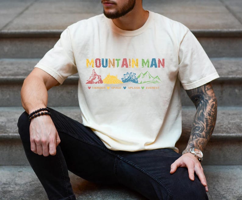 May include: A person wearing a light beige t-shirt with the text "Mountain Man" and a graphic of four mountains with the words "Thrill", "Space", "Splash", and "Everest" underneath. The person is wearing black jeans and a watch on their left wrist.