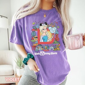 Może przedstawiać: Fioletowy T-shirt z grafiką postaci Disneya i tekstem "Walt Disney World". Grafika przedstawia kresk&oacute;wkową postać Elsy z Krainy Lodu, Myszki Miki i innych postaci Disneya.
