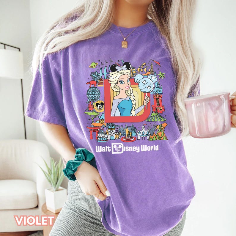 Może przedstawiać: Fioletowy T-shirt z grafiką postaci Disneya i tekstem "Walt Disney World". Grafika przedstawia kresk&oacute;wkową postać Elsy z Krainy Lodu, Myszki Miki i innych postaci Disneya.