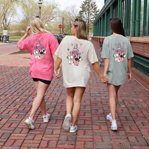 May include: Three women walking on a brick path, each wearing a different colored oversized t-shirt. The shirts are pink, cream, and sage green, all featuring a colorful graphic of a castle and characters on the back.