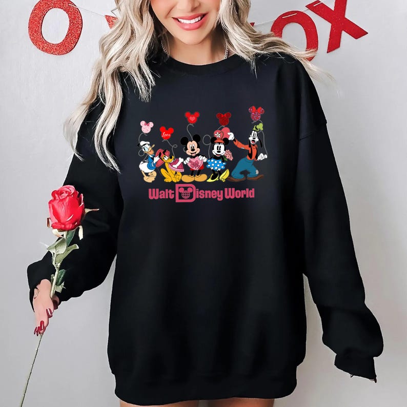 K&ouml;nnte beinhalten: Schwarzer Pullover mit einem Aufdruck von Mickey Mouse, Minnie Mouse, Donald Duck, Goofy und Pluto, die Herzen halten. Der Text "Walt Disney World" ist unter dem Aufdruck gedruckt.