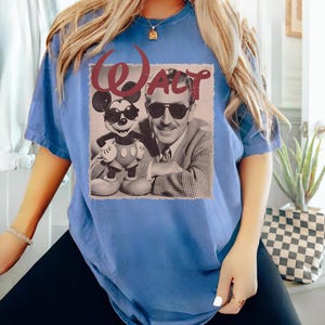 Op de afbeelding: Een blauw t-shirt met korte mouwen en een grafische print van Walt Disney en Mickey Mouse die beide een zonnebril dragen. De afbeelding staat op een lichte vierkante achtergrond met het woord WALT in grote, donkerrode cursieve letters erboven.