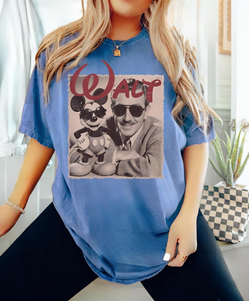 Op de afbeelding: Een blauw t-shirt met korte mouwen en een grafische print van Walt Disney en Mickey Mouse die beide een zonnebril dragen. De afbeelding staat op een lichte vierkante achtergrond met het woord WALT in grote, donkerrode cursieve letters erboven.