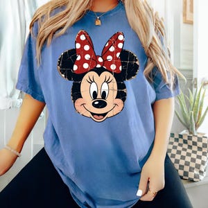 May include: A blue short-sleeved t-shirt featuring a distressed graphic print of Minnie Mouse on the front. The character design includes her signature black ears and a red bow with white polka dots, rendered with a weathered, cracked texture.