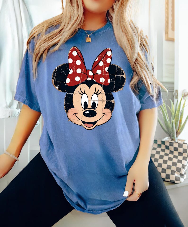 May include: A blue short-sleeved t-shirt featuring a distressed graphic print of Minnie Mouse on the front. The character design includes her signature black ears and a red bow with white polka dots, rendered with a weathered, cracked texture.