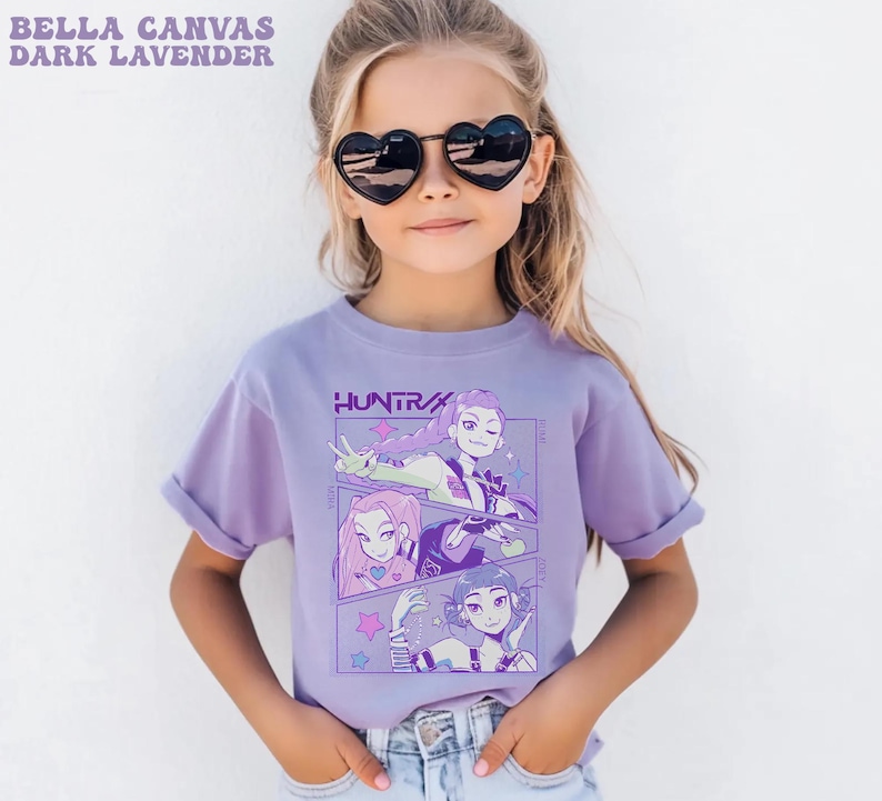 Peut inclure: T-shirt lavande fonc&eacute; avec un motif de trois personnages de style anime. Le t-shirt a des manches retrouss&eacute;es et le texte "HUNTRA" est visible. Le texte "BELLA CANVAS DARK LAVENDER" est visible en haut &agrave; gauche.