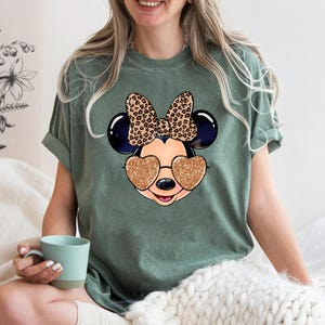 以下が含まれることがあります： キラキラしたハート型のサングラスをかけたマウスの漫画の画像がプリントされた緑色のTシャツ。マウスはレオパード柄の黒いリボンをしています。