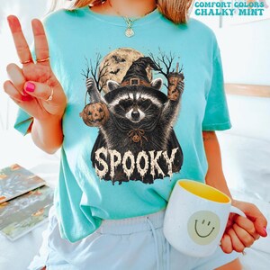 Pu&ograve; includere: Una t-shirt a maniche corte color verde menta gessato con la grafica di un procione che indossa un cappello da strega e un mantello, mentre tiene in mano un secchiello a forma di zucca e una bevanda. Include luna piena, alberi e la scritta "SPOOKY".
